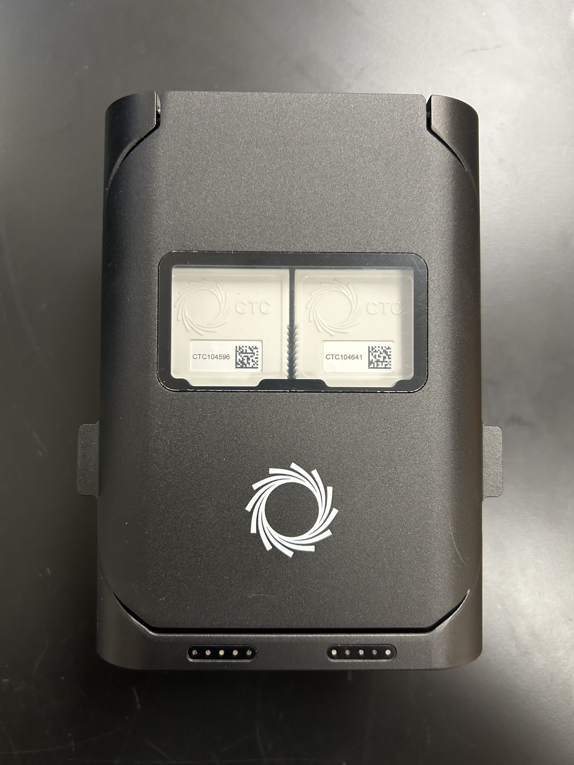 IMG_7062 A black rectangular electronic device with two visible labeled chip slots under a clear window, featuring a white circular logo on the front, placed on a dark surface.