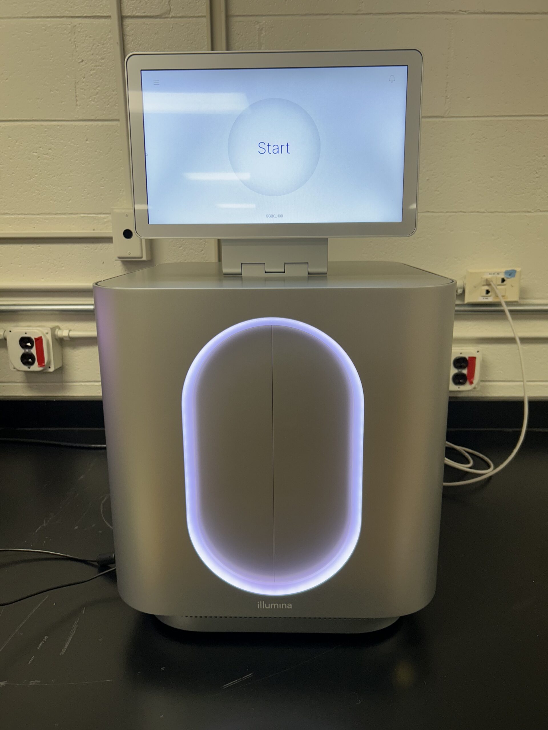 A DNA sequencing machine with a touchscreen displaying a Start button, positioned in a lab with visible power outlets and cables on the wall.